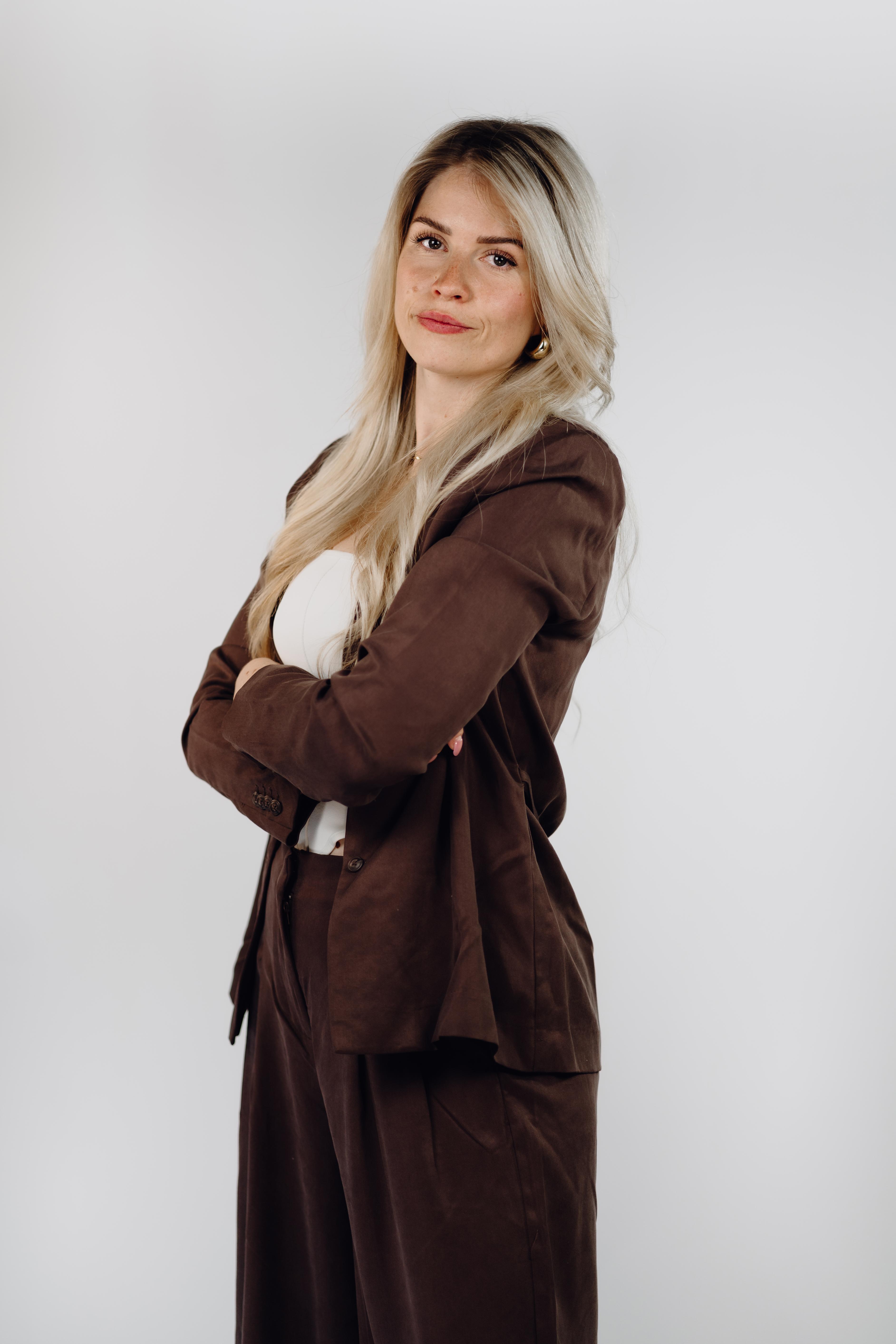 Professional headshot of a woman with long blonde hair, standing at an angle with arms crossed, wearing a brown blazer and matching wide-leg trousers.