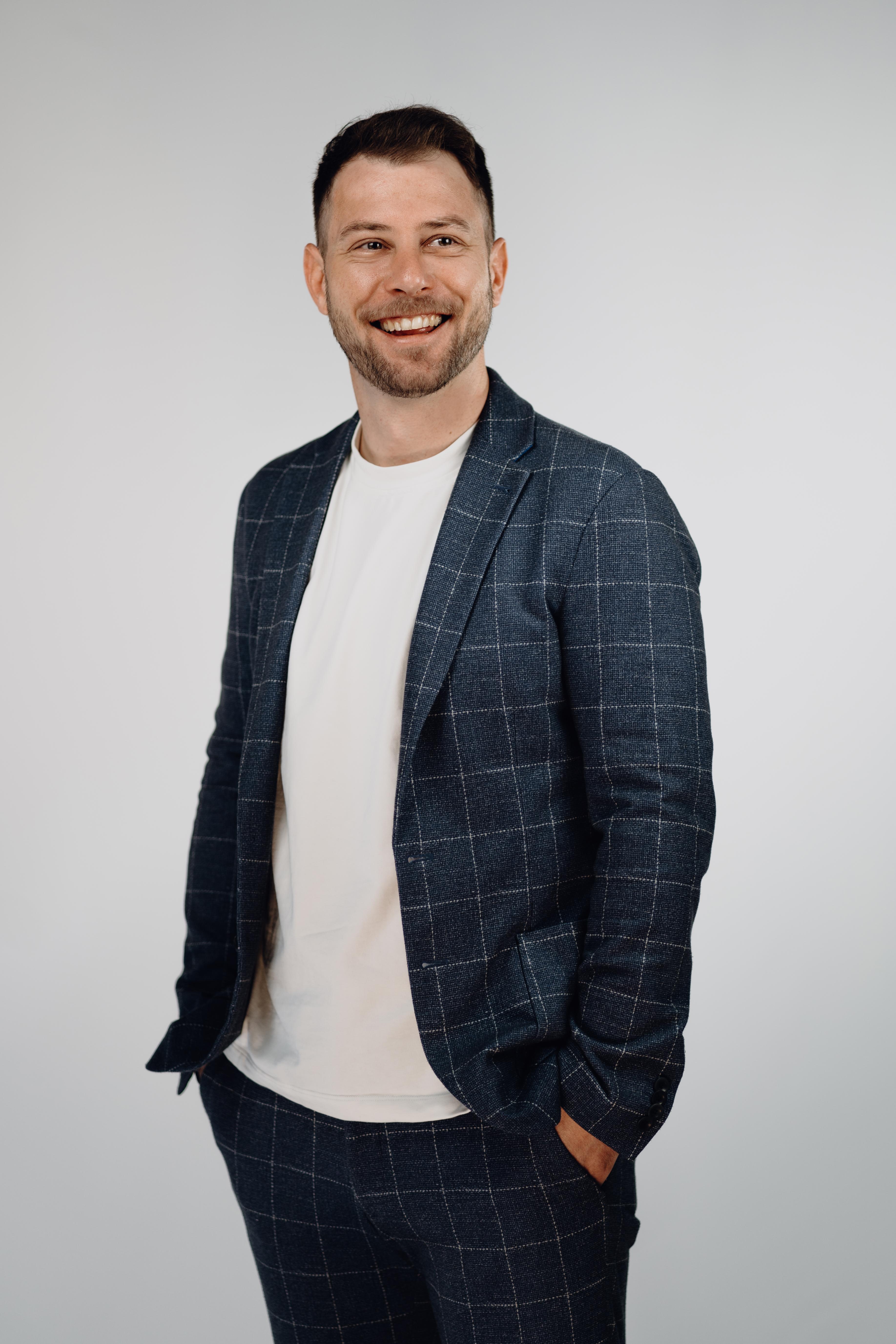 Professional headshot of a man with short dark hair and a beard, smiling broadly, wearing a navy windowpane-check blazer over a white T-shirt with one hand in his pocket.