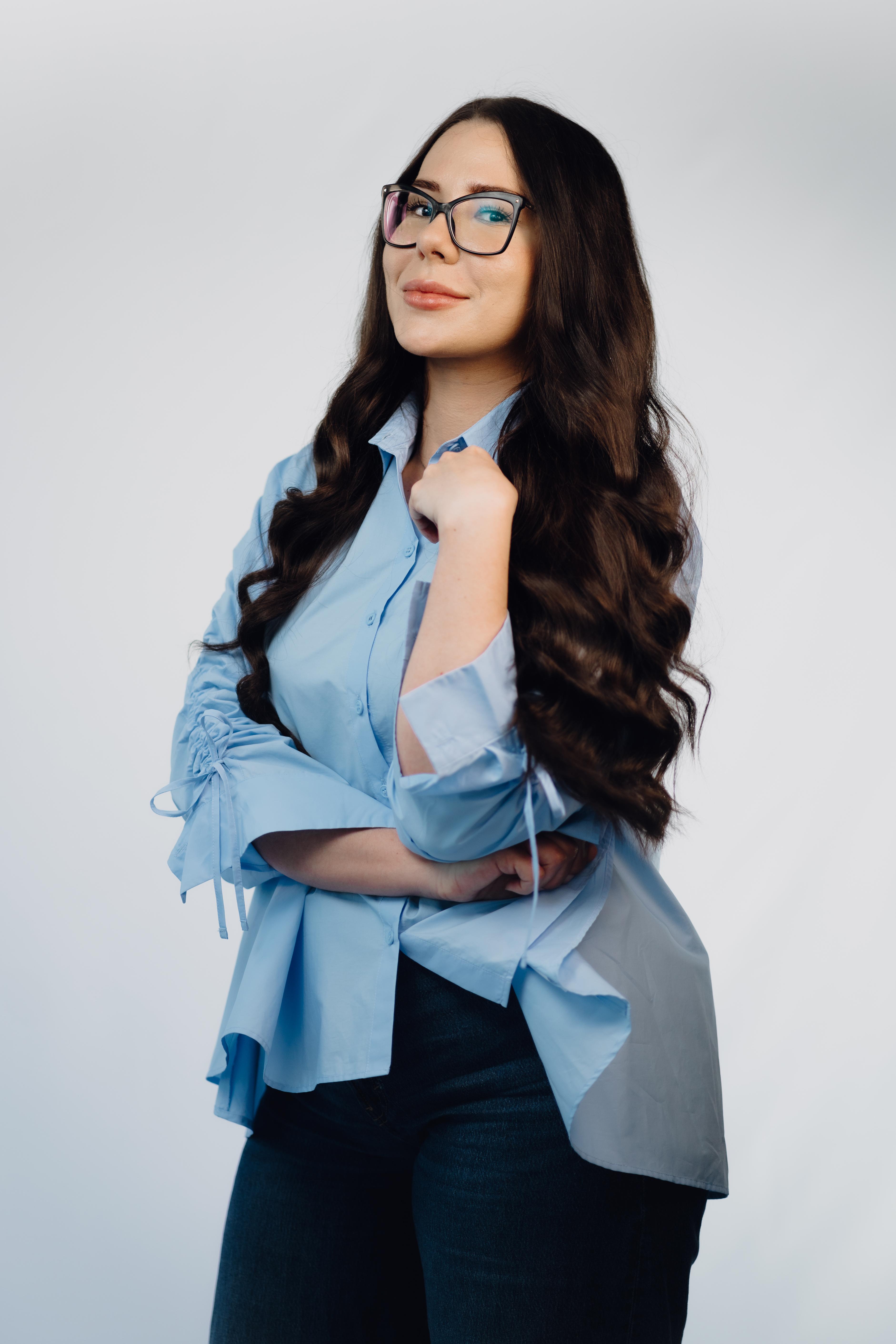 Professional headshot of a woman with long dark wavy hair and glasses, wearing a light blue blouse and dark jeans, arms crossed, smiling slightly at the camera.