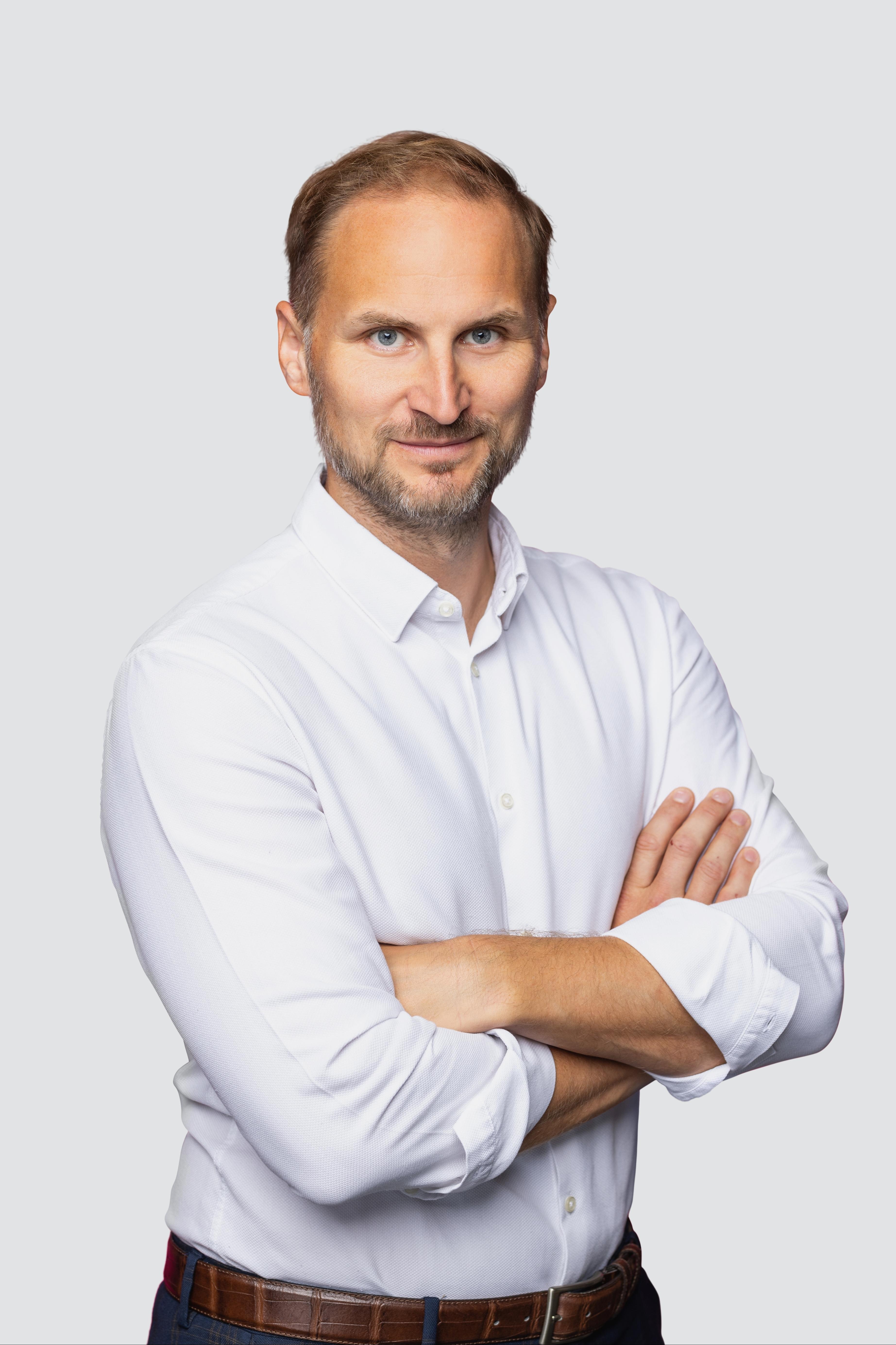 Professional headshot of a man with short reddish-brown hair and a light beard, wearing a white button-down shirt with sleeves rolled up, standing with arms crossed and looking at the camera.