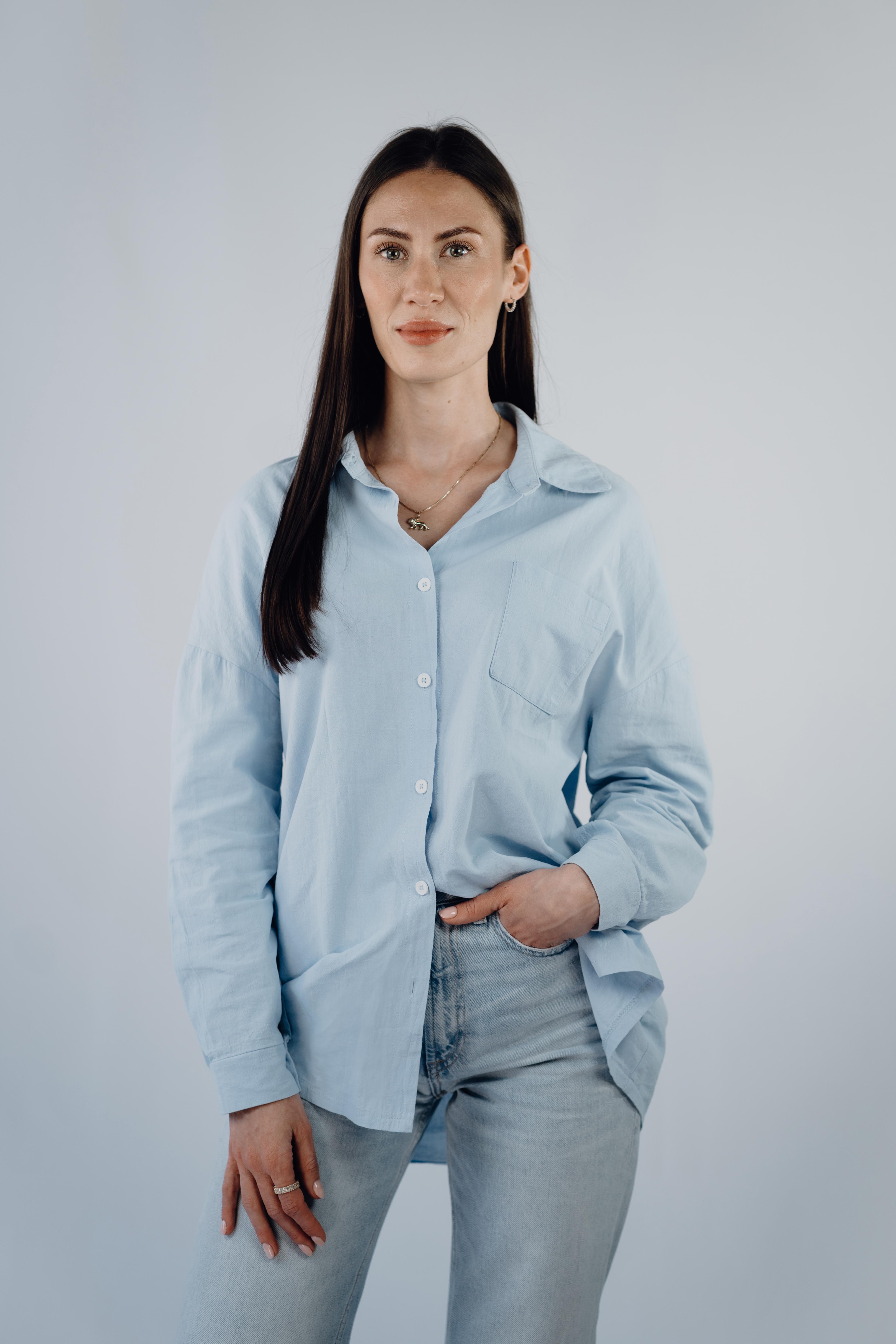 Professional headshot of a woman with long straight dark hair, wearing a pale blue button-down shirt and jeans, one hand in her pocket, looking at the camera with a neutral expression.