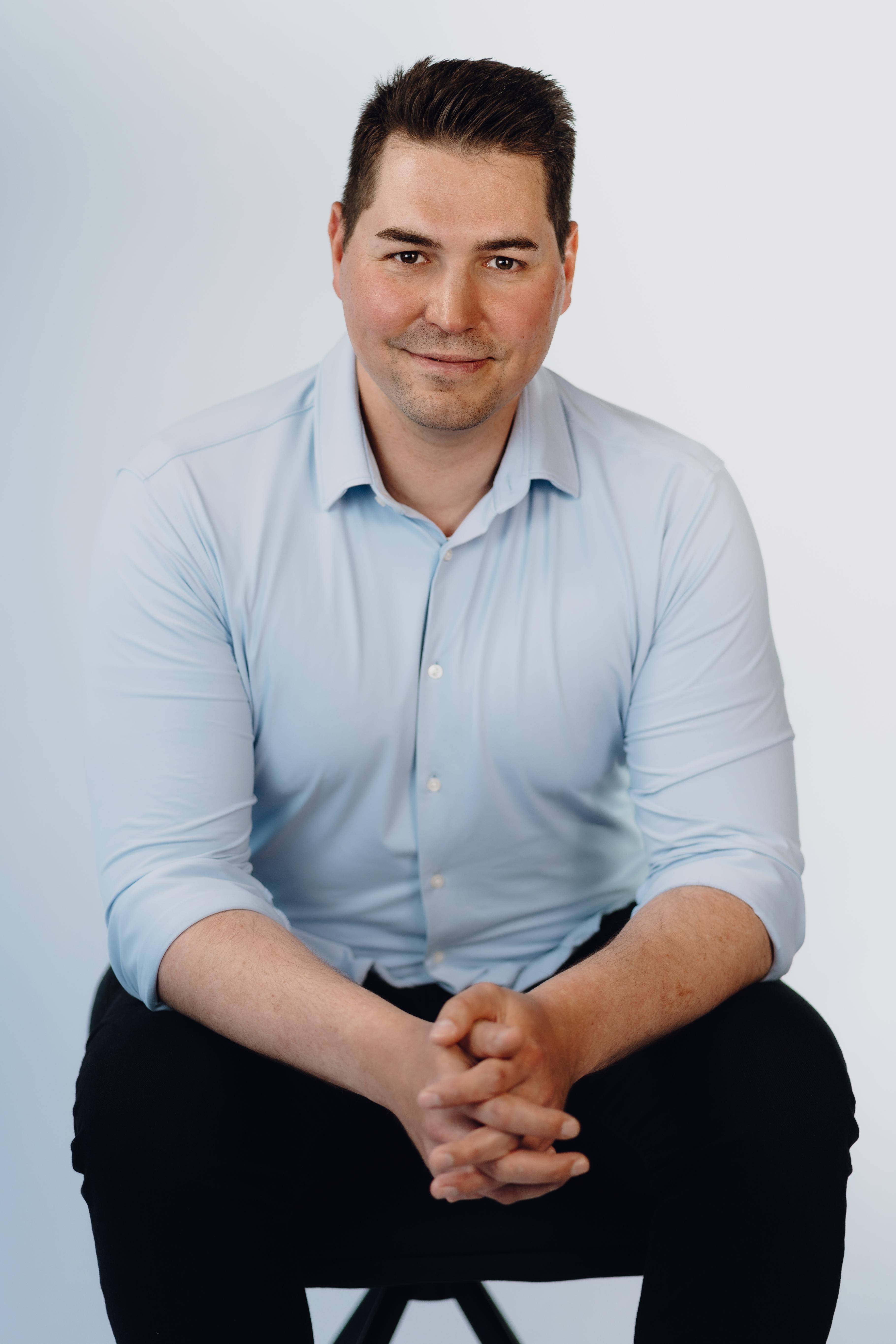 Professional headshot of a man with short dark hair, seated on a stool wearing a light blue button-down shirt, hands clasped over his knee, smiling gently at the camera.