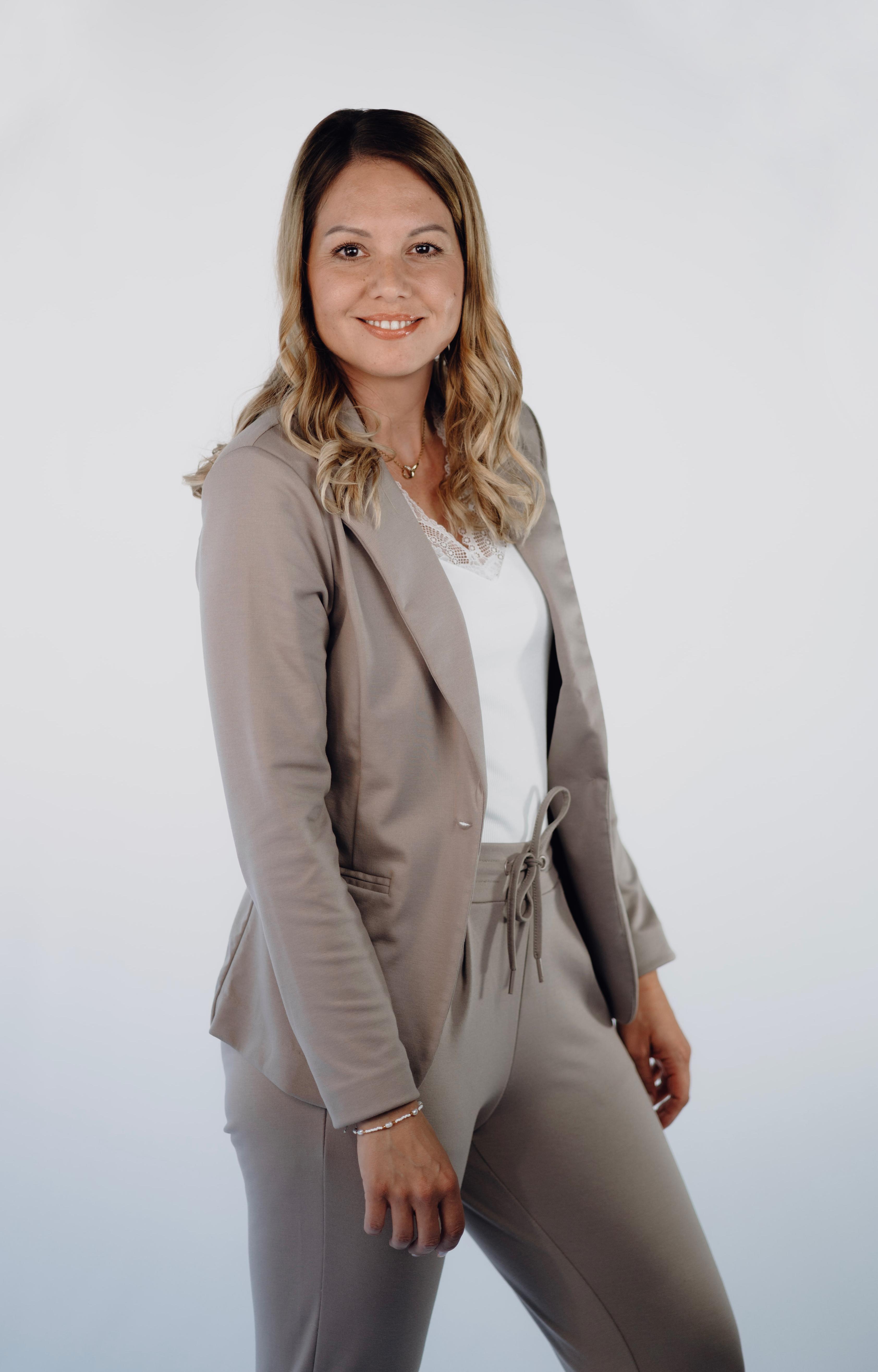 Professional headshot of a woman with wavy blonde hair and highlights, smiling, wearing a taupe blazer and matching trousers over a white top.