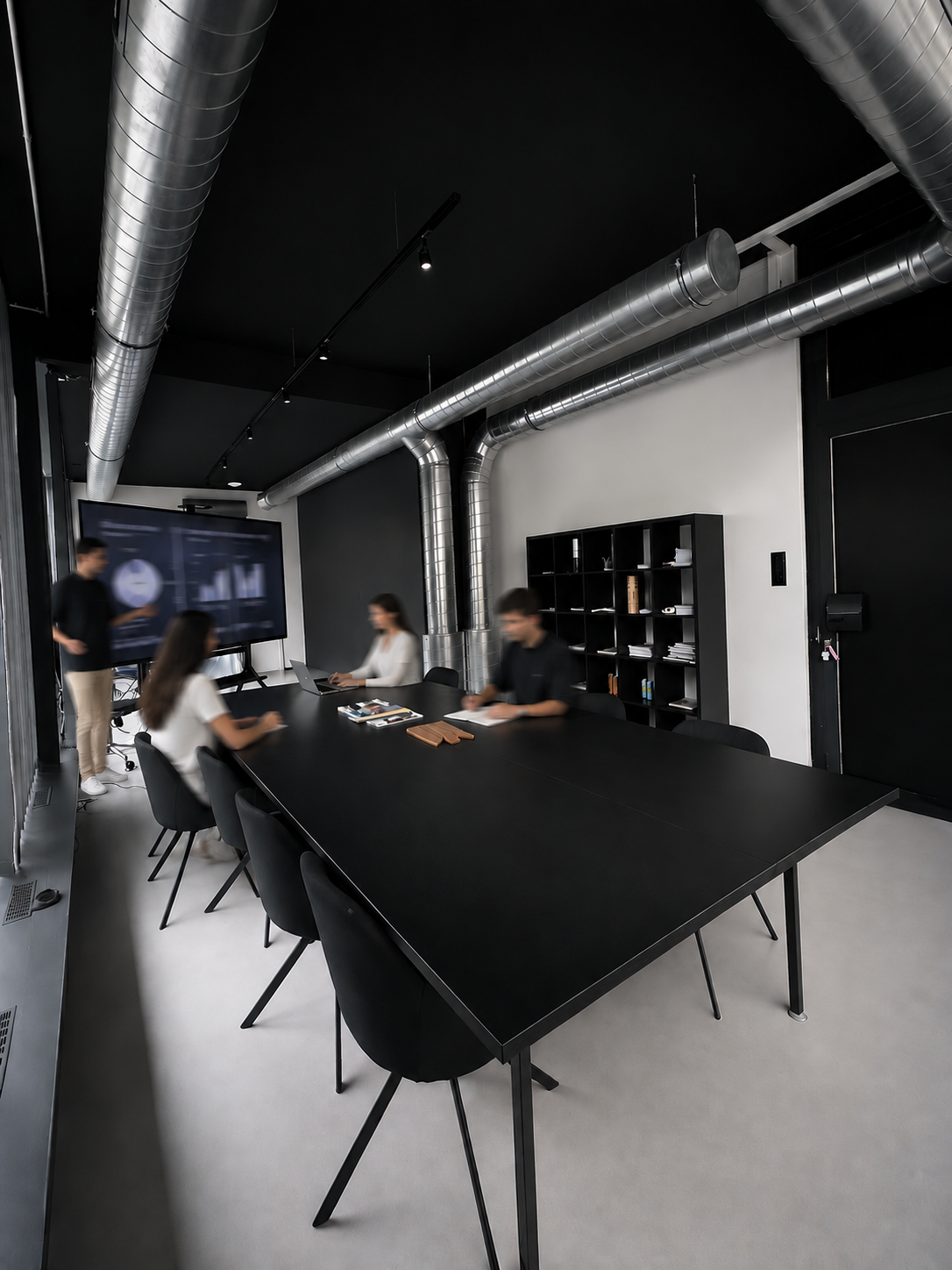 Modern meeting room with a black conference table and chairs, exposed silver ductwork along a black ceiling, and four colleagues attending a presentation, one standing at a wall-mounted screen showing charts.