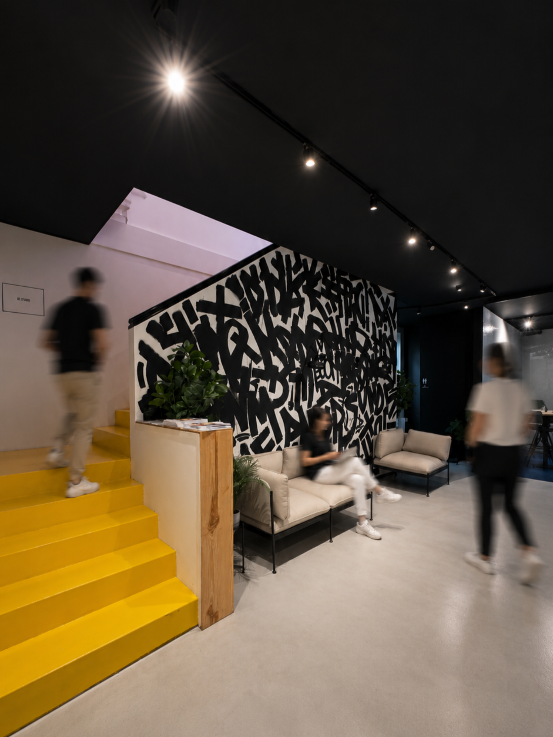 Office lobby with bright yellow stairs and a black-and-white graffiti-style accent wall, one person sitting on a beige sofa and two others walking past in motion blur, under warm spotlighting.