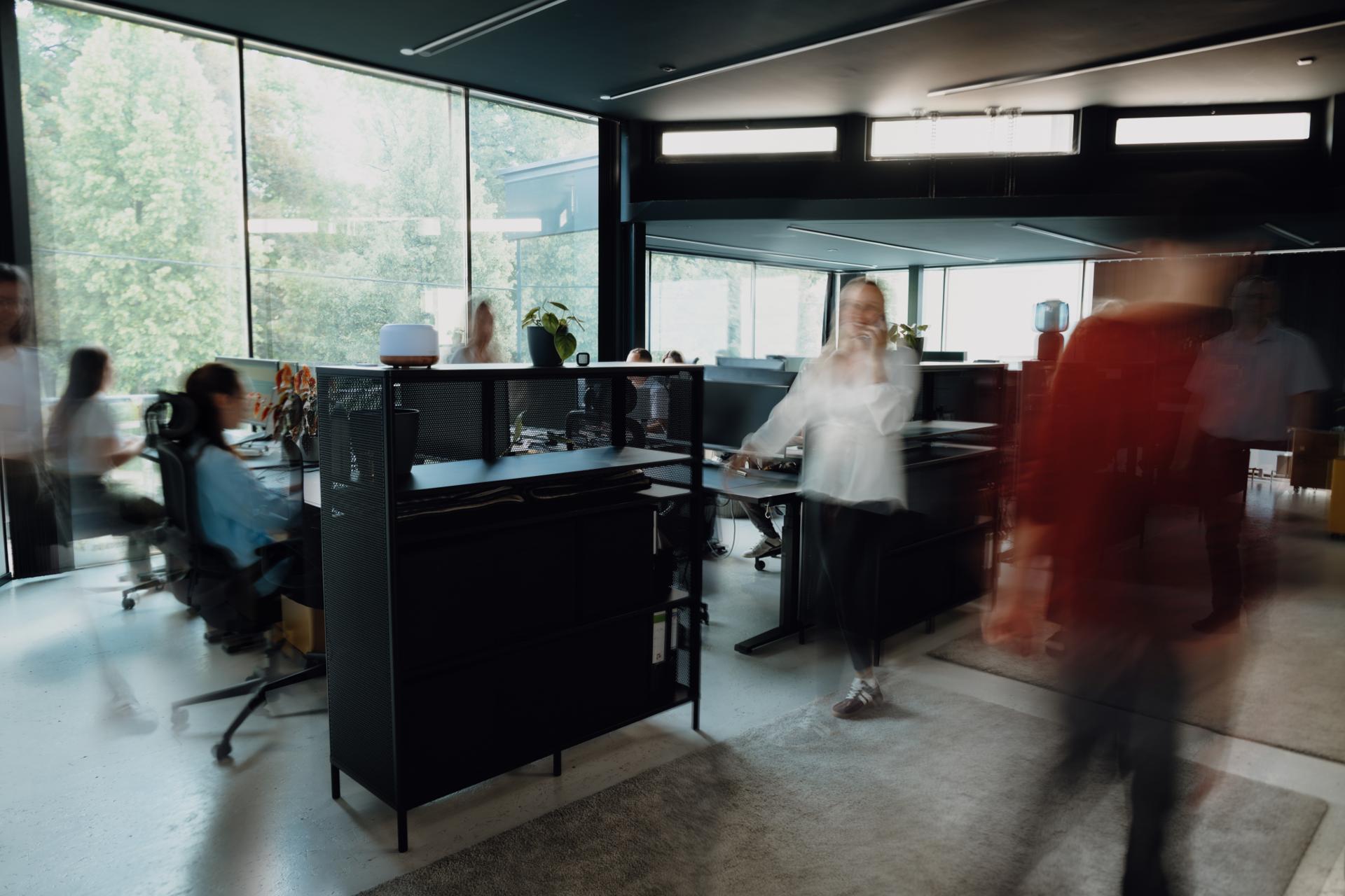 Blurred, fast-paced open office with employees moving between standing desks and workstations, floor-to-ceiling windows showing greenery outside, and a dark ceiling with exposed beams.