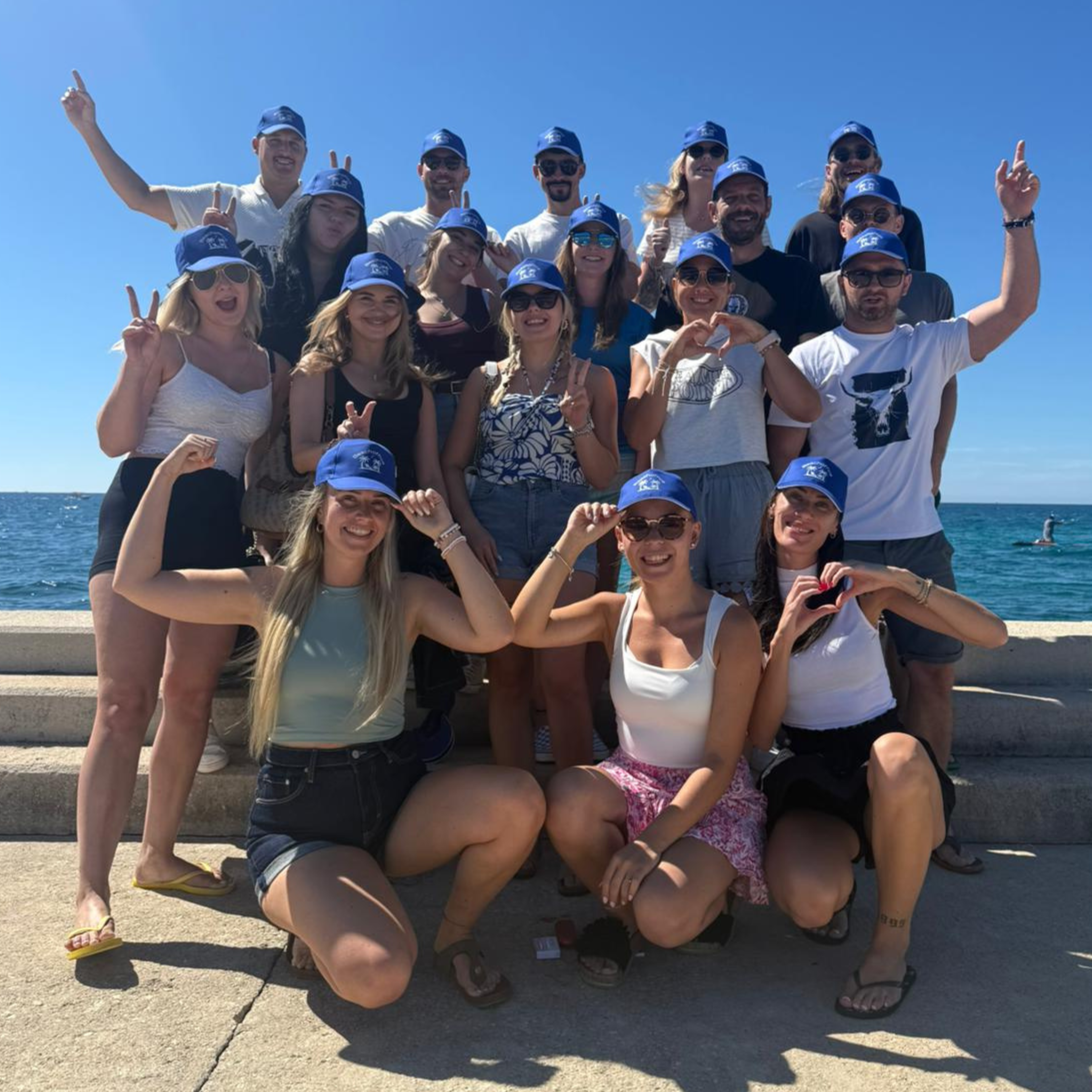 Group of about eighteen people wearing matching navy blue caps posing together on a stone pier by the sea, most making peace signs or raising fists, with several kneeling in front and the rest standing behind them under a clear blue sky.