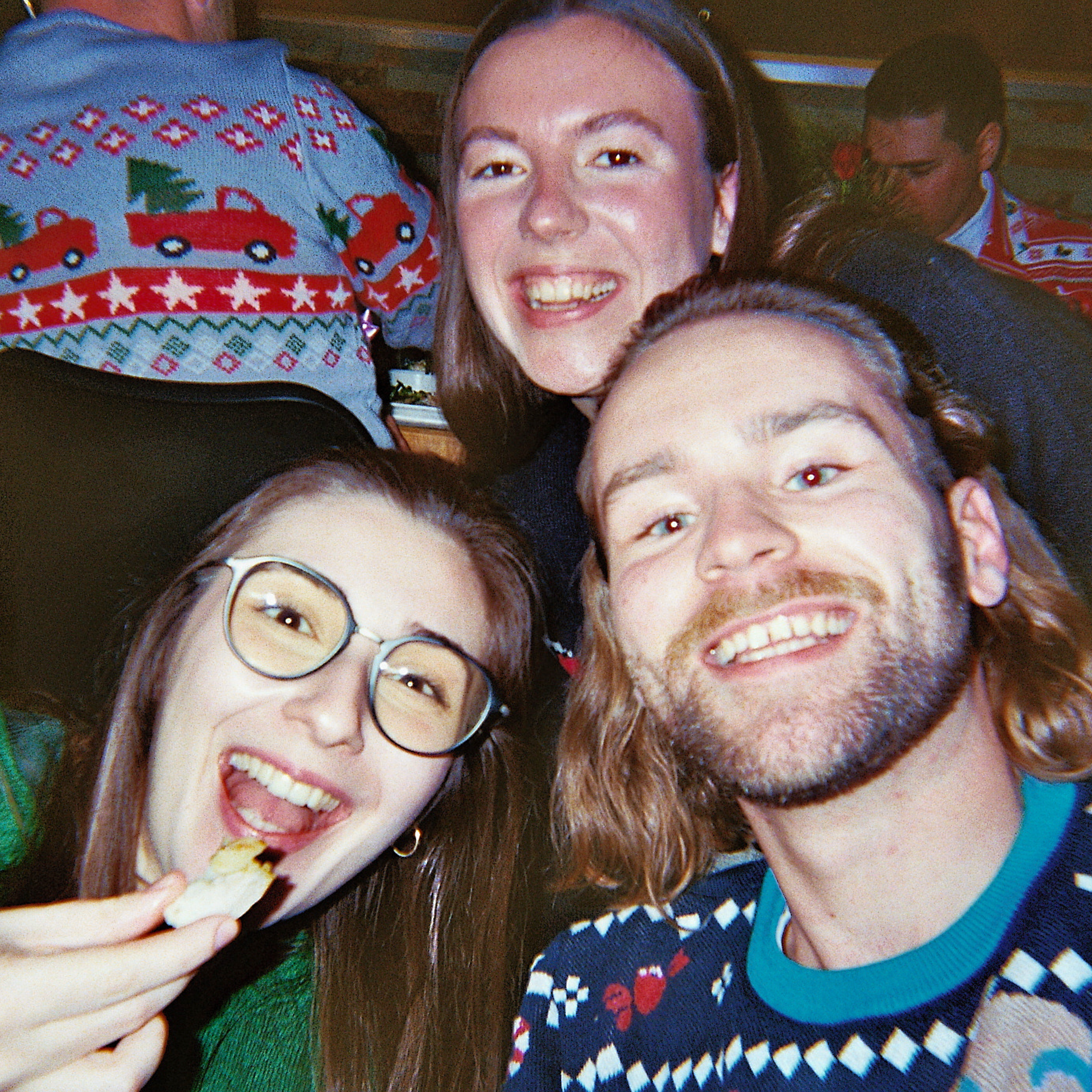 Close-up selfie of three friends smiling at a festive party, two wearing Christmas-patterned sweaters and one eating a snack, with a blurred crowd visible in the background.