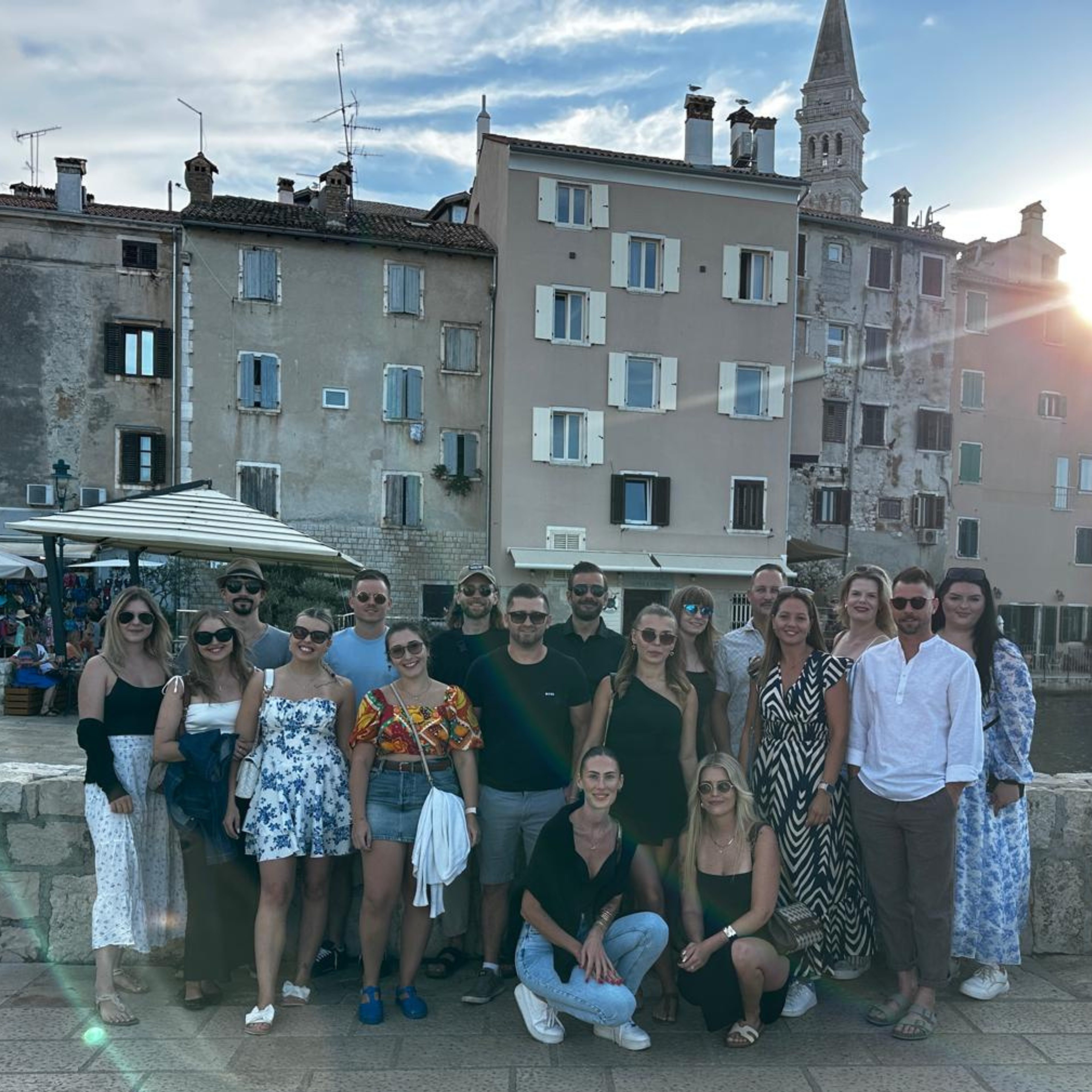 Large group of about seventeen people posing together at sunset on a stone waterfront promenade in front of a row of pastel-coloured European buildings and a bell tower, with some kneeling in front and others standing behind.