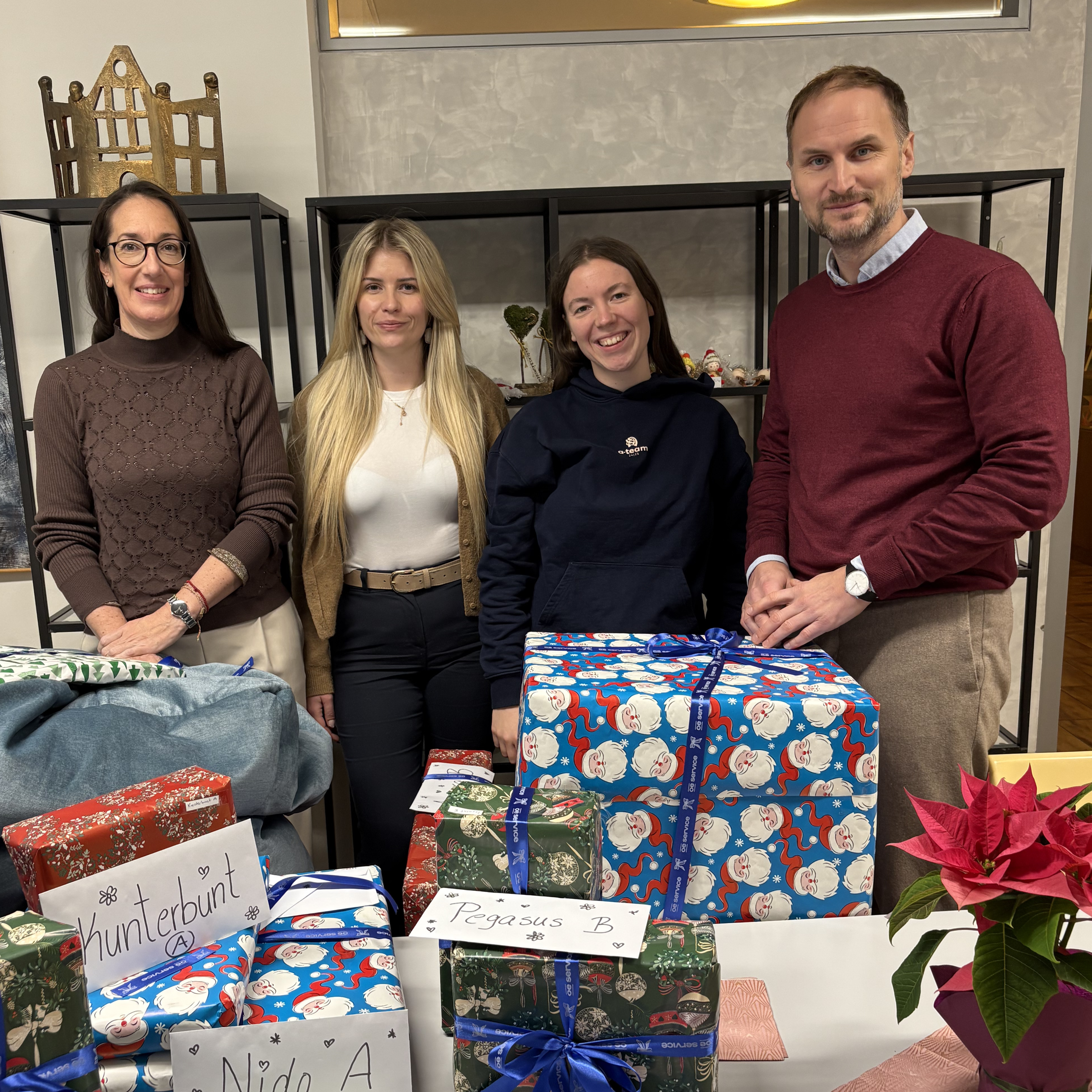 Four colleagues standing behind a table piled with wrapped Christmas gifts labelled with names, smiling at the camera in an office with glass display cabinets and a poinsettia plant in the corner.