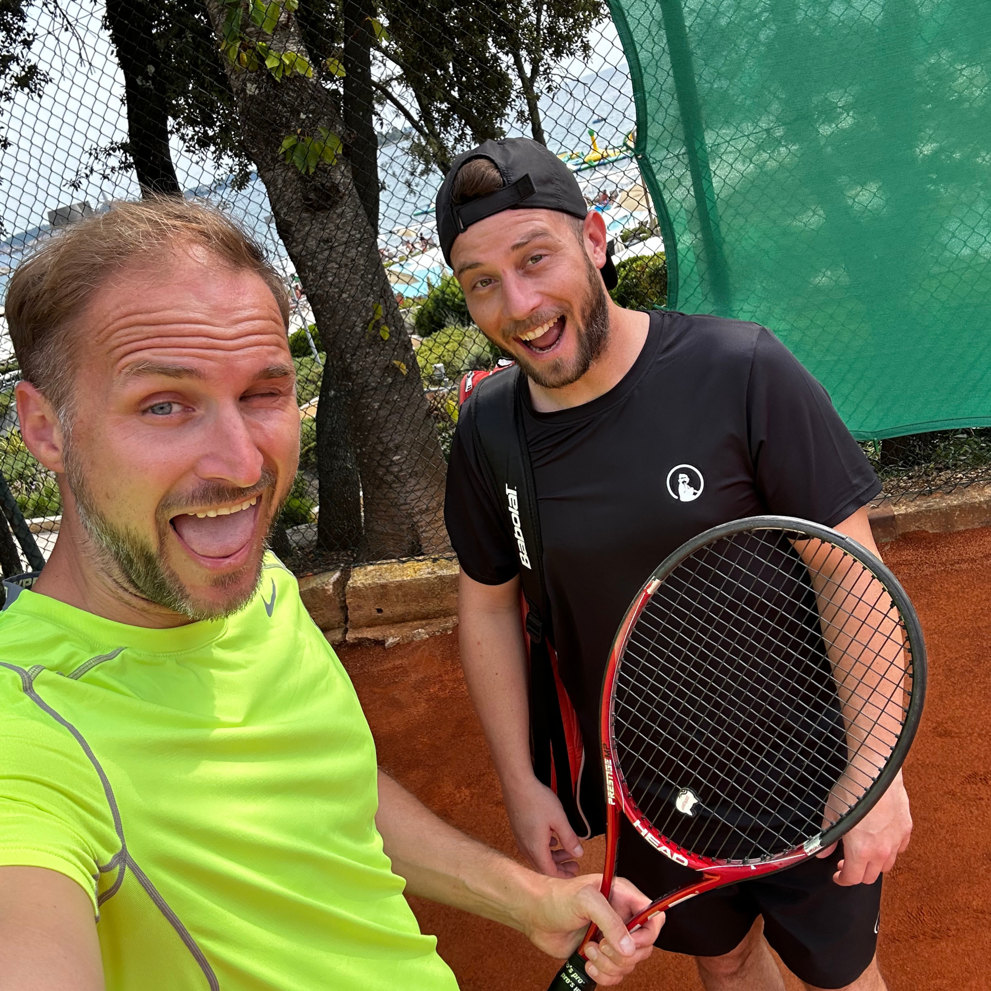 Two men taking a selfie on a red clay tennis court, one in a bright yellow-green shirt and the other in black holding a tennis racket, both smiling with trees and a green fence in the background.